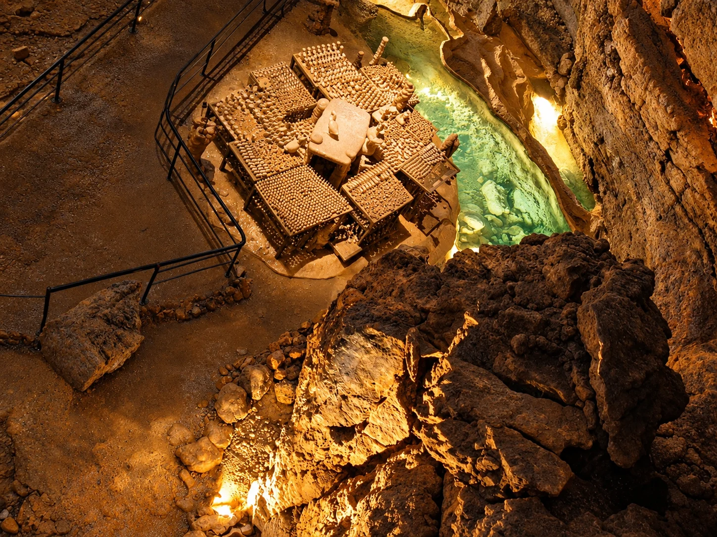An elevated view shows trays of objects beside an illuminated pool and railed paths inside the Gouffre de Proumeyssac.