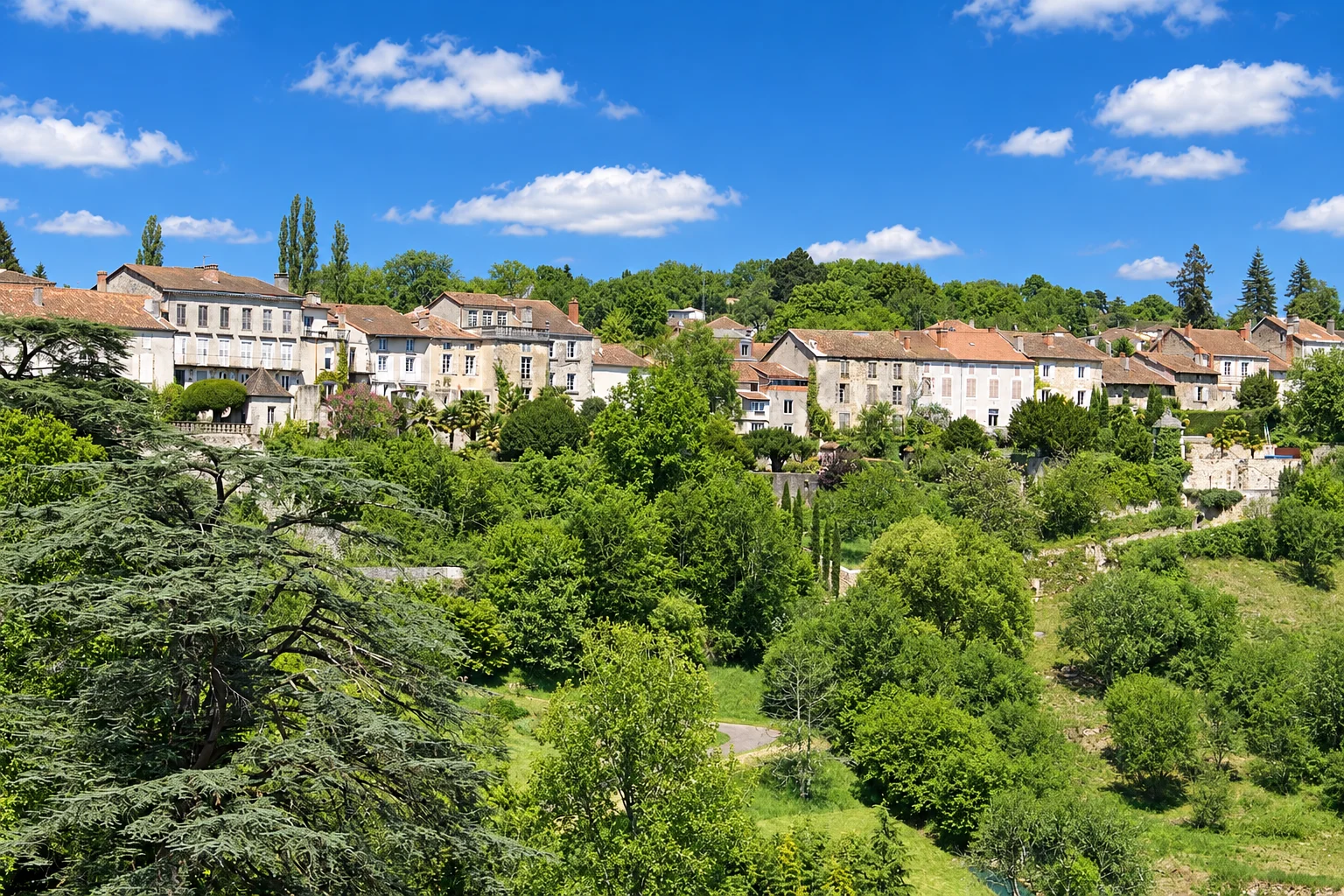 Houses in Nontron overlook terraced gardens and a green valley beneath a partly cloudy sky.