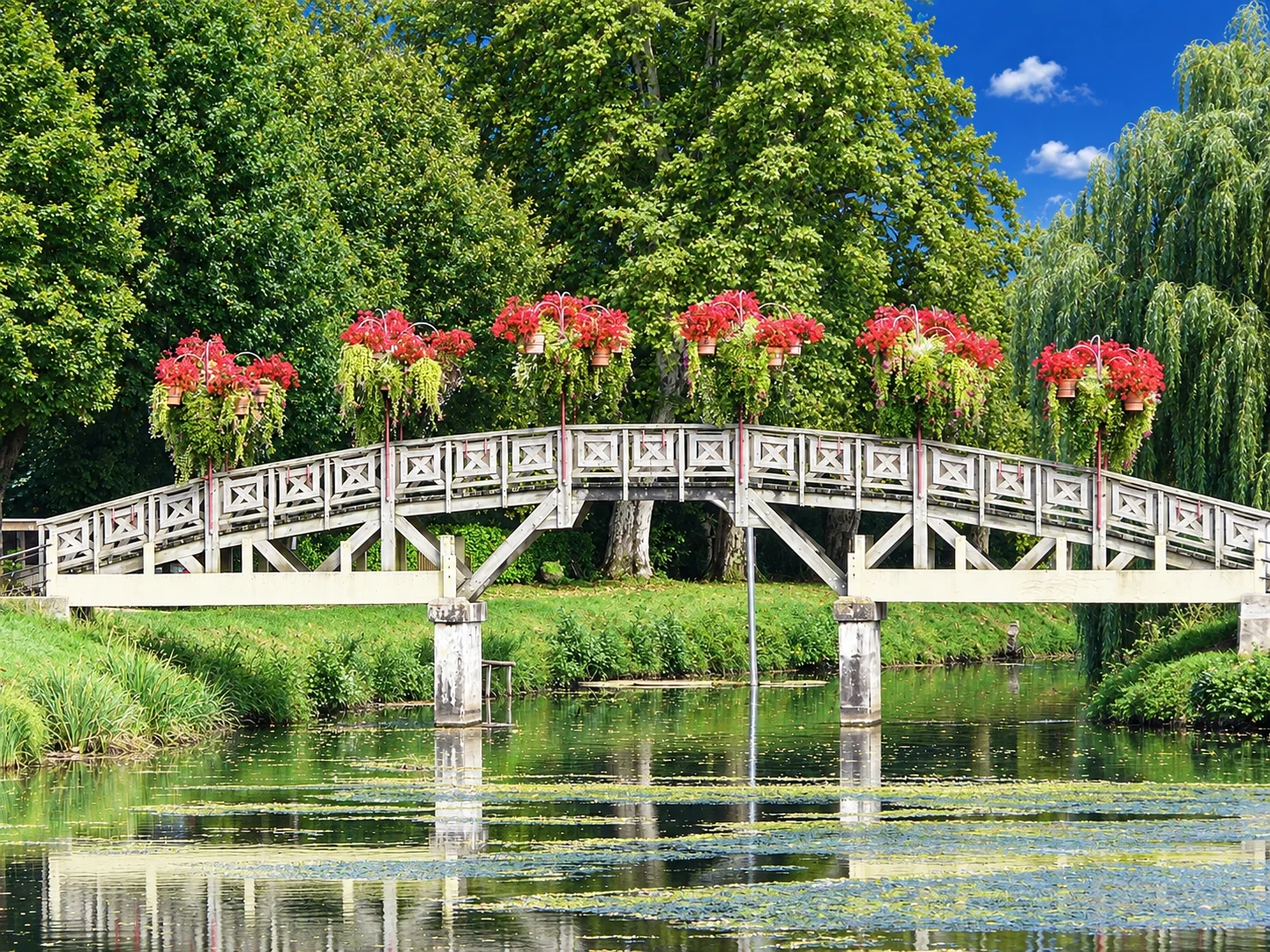 Hanging baskets of red flowers line a wooden footbridge reflected in the water at Lalinde.