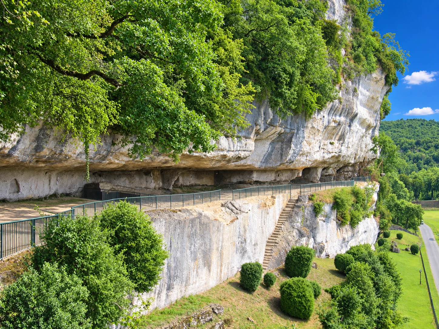 Visitors follow the limestone cliff terrace at La Roque Saint-Christophe, above a road and the wooded Vézère valley.