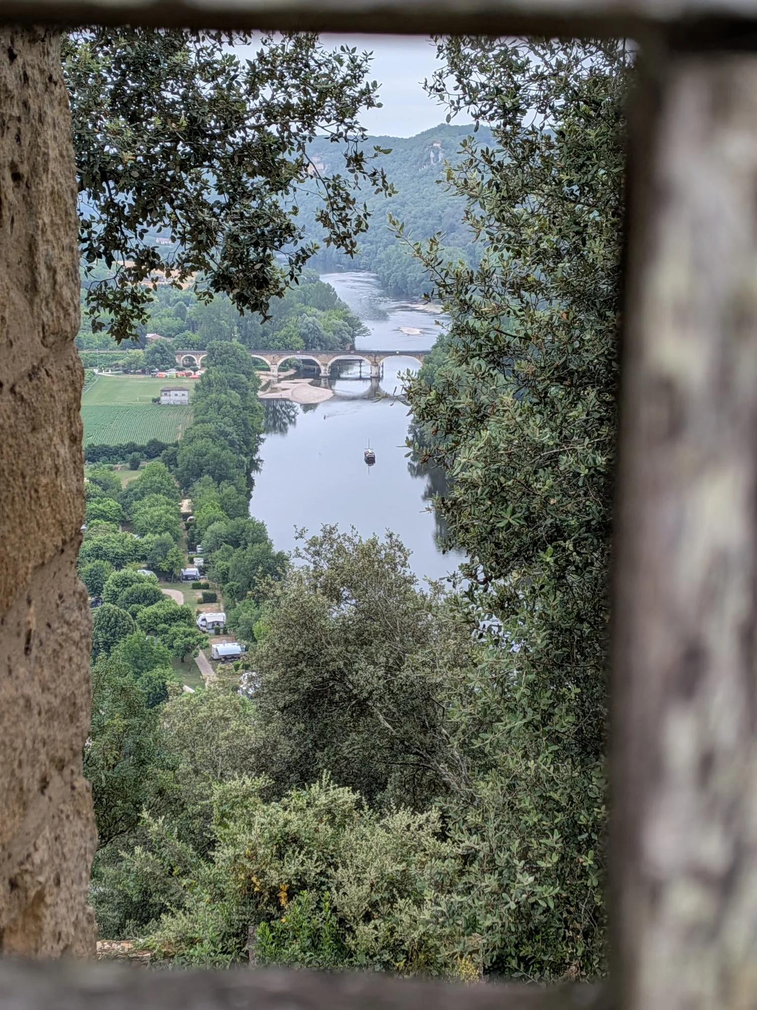 View through a stone window at Château de Beynac down the Dordogne towards an arched stone bridge and a gabarre