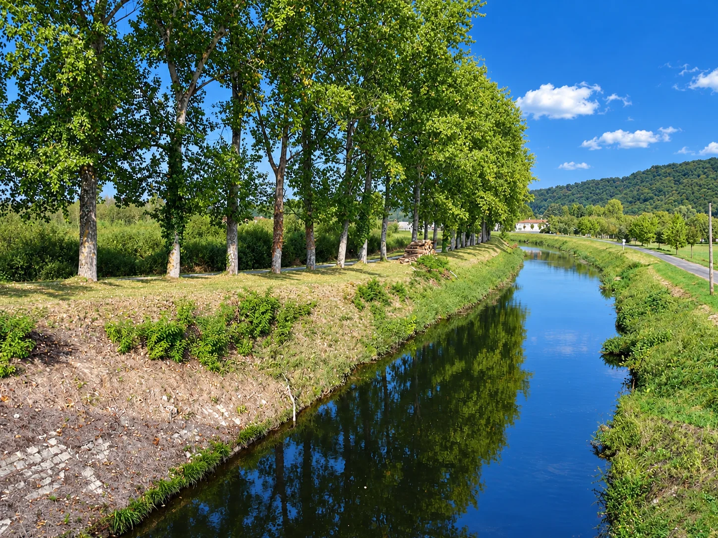 A row of trees follows the Lalinde canal, with reflections in the water and wooded hills beyond.